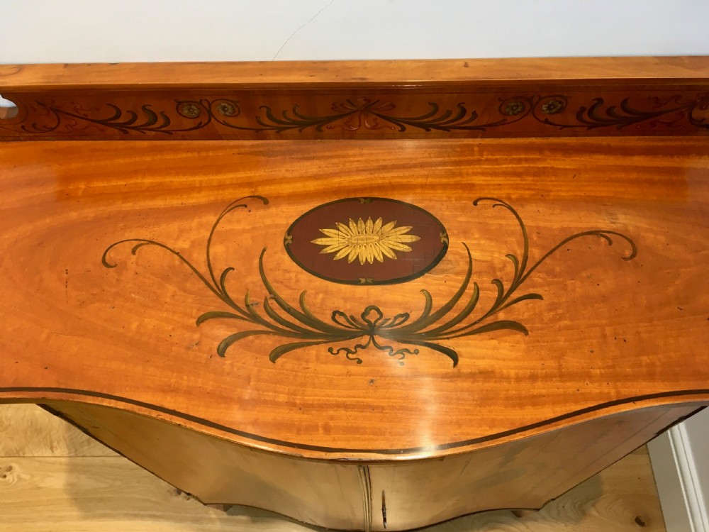 A satinwood side cabinet featuring a serpentine front, twin cupboard doors decorated with hand-painted Neoclassical urns and floral swags, supported by tapered legs. View XV