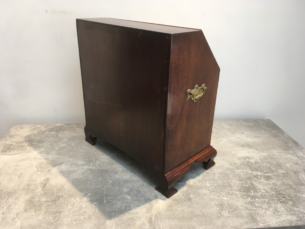 A miniature dark mahogany fall front bureau featuring tiered drawers, side carrying handles, brass hardware, and ogee bracket feet. View V