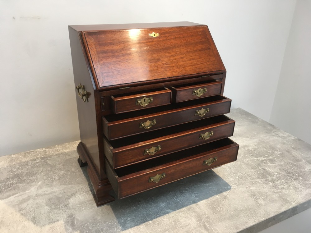 A miniature dark mahogany fall front bureau featuring tiered drawers, side carrying handles, brass hardware, and ogee bracket feet. View VI