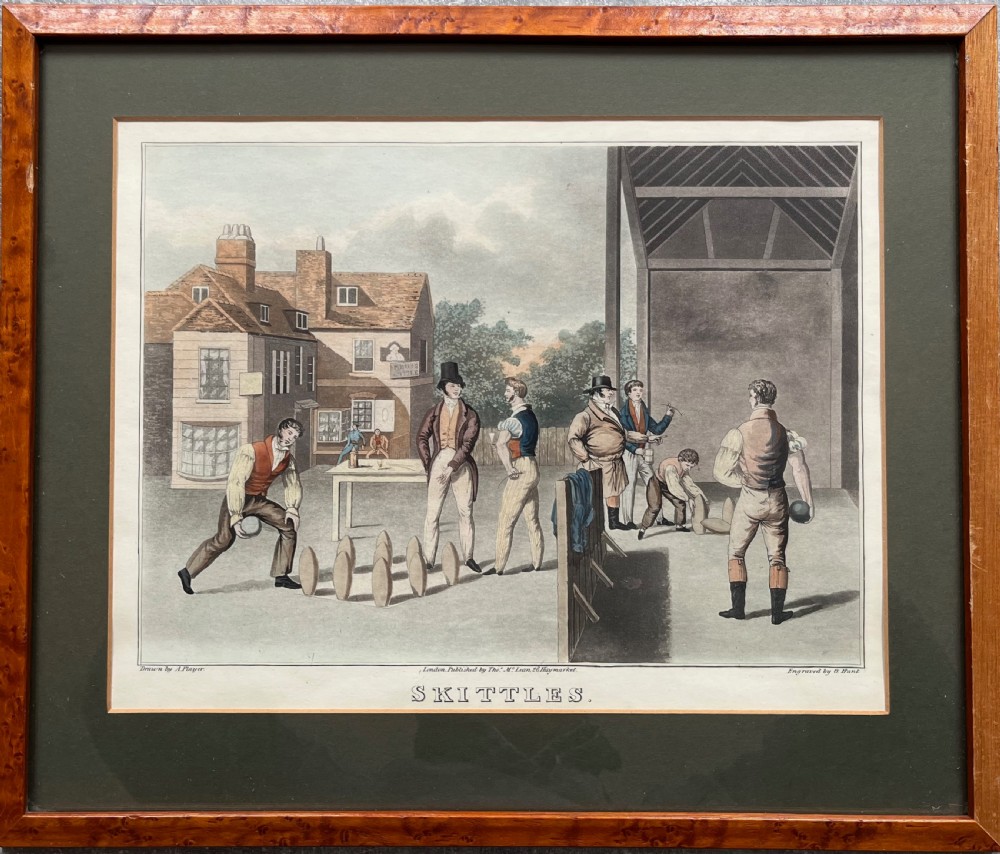 A framed antique hand-coloured engraving depicting gentlemen playing skittles outside an English pub, set within a dark green mount and a warm wood frame.