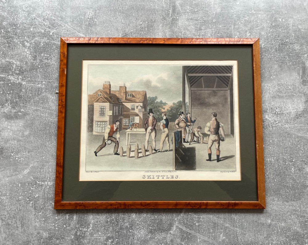 A framed antique hand-coloured engraving depicting gentlemen playing skittles outside an English pub, set within a dark green mount and a warm wood frame. View VII