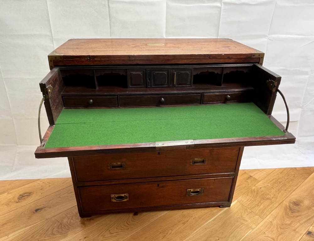 A teak campaign secretaire chest featuring brass corner brackets, flush brass drawer handles, and an open fall-front writing surface lined with green felt.