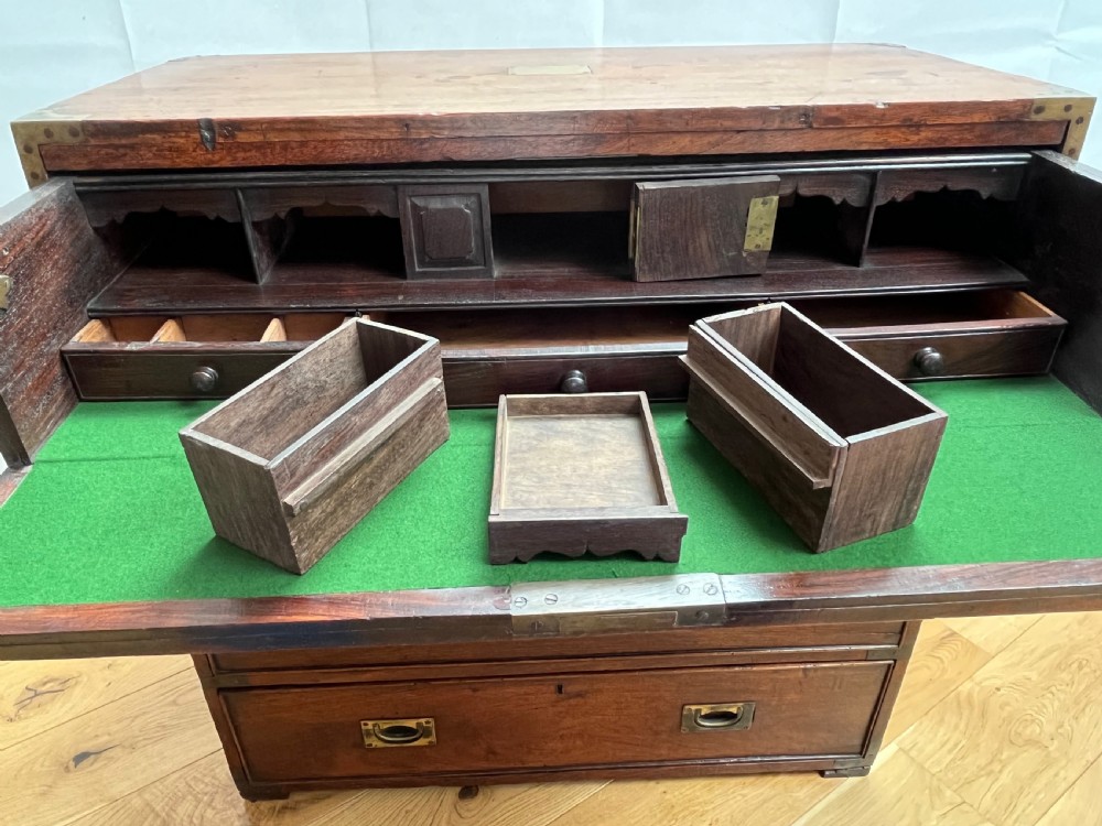 A teak campaign secretaire chest featuring brass corner brackets, flush brass drawer handles, and an open fall-front writing surface lined with green felt. View VI