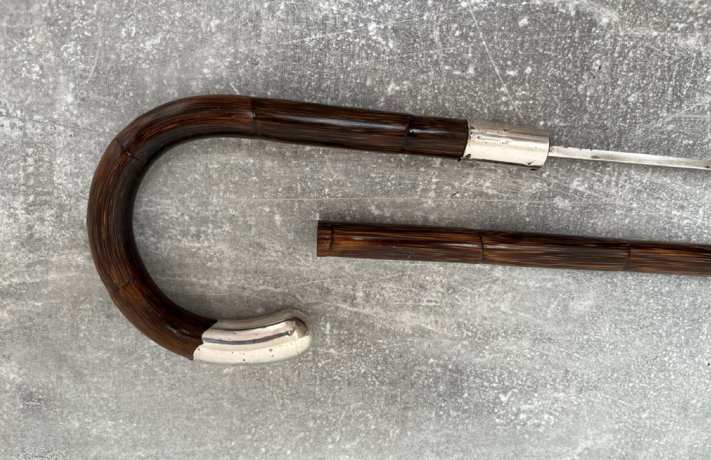 A dark brown malacca cane walking stick featuring a curved crook handle, a silver end cap, a silver collar, and a brass ferrule at the tip. View IV