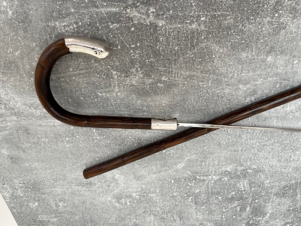 A dark brown malacca cane walking stick featuring a curved crook handle, a silver end cap, a silver collar, and a brass ferrule at the tip. View VI