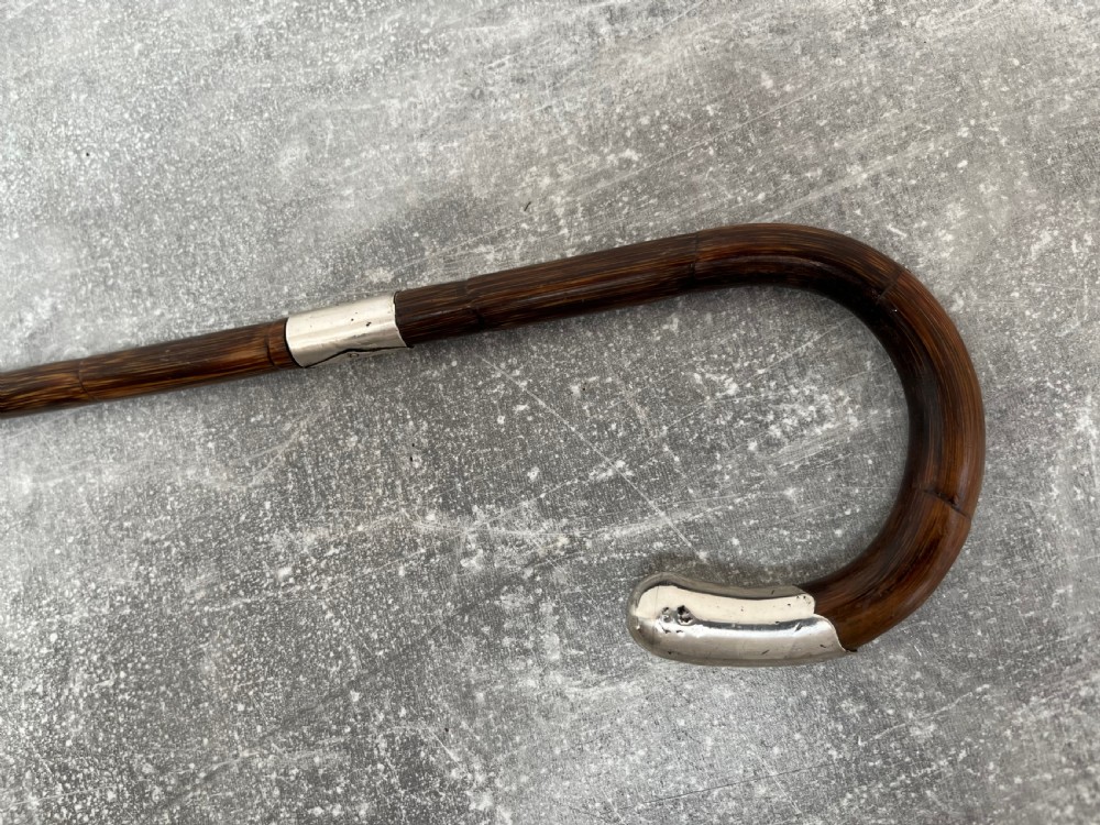 A dark brown malacca cane walking stick featuring a curved crook handle, a silver end cap, a silver collar, and a brass ferrule at the tip. View X