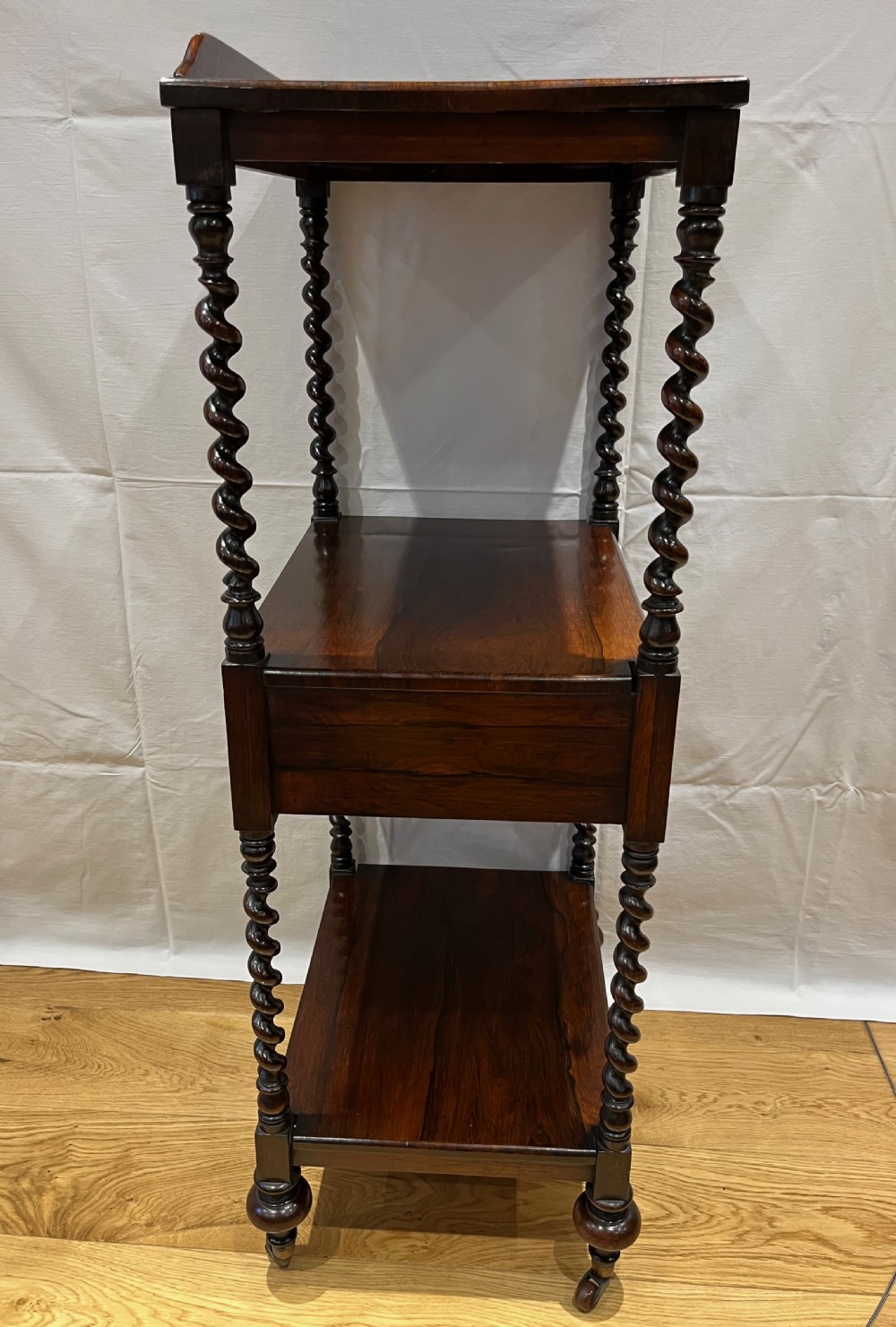 A mid-Victorian dark rosewood whatnot etagere featuring three open tiers, barley twist pillar supports, a central drawer, a gallery back, and turned feet. View XI