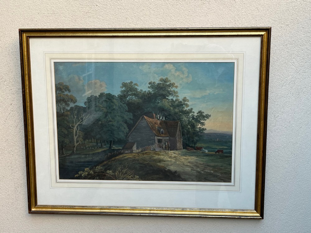 A framed 19th-century watercolour painting featuring a rustic cottage nestled among lush trees with livestock in a rolling landscape, presented in a gilt frame.