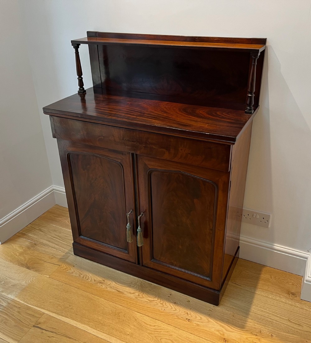 A Regency flame mahogany chiffonier cabinet featuring a top display shelf supported by turned columns over a frieze drawer and two panelled doors.