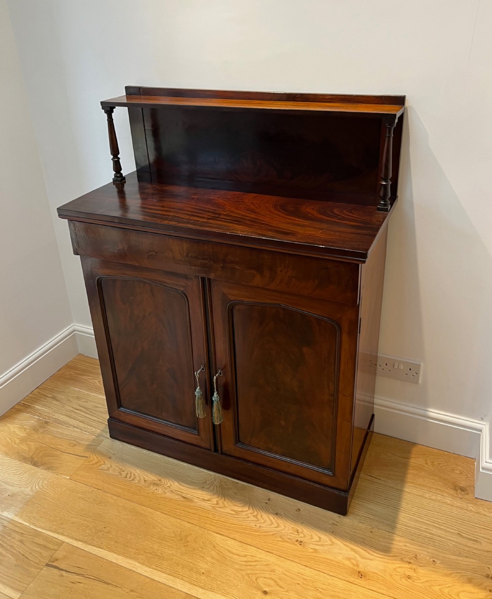 A Regency flame mahogany chiffonier cabinet featuring a top display shelf supported by turned columns over a frieze drawer and two panelled doors. View IV
