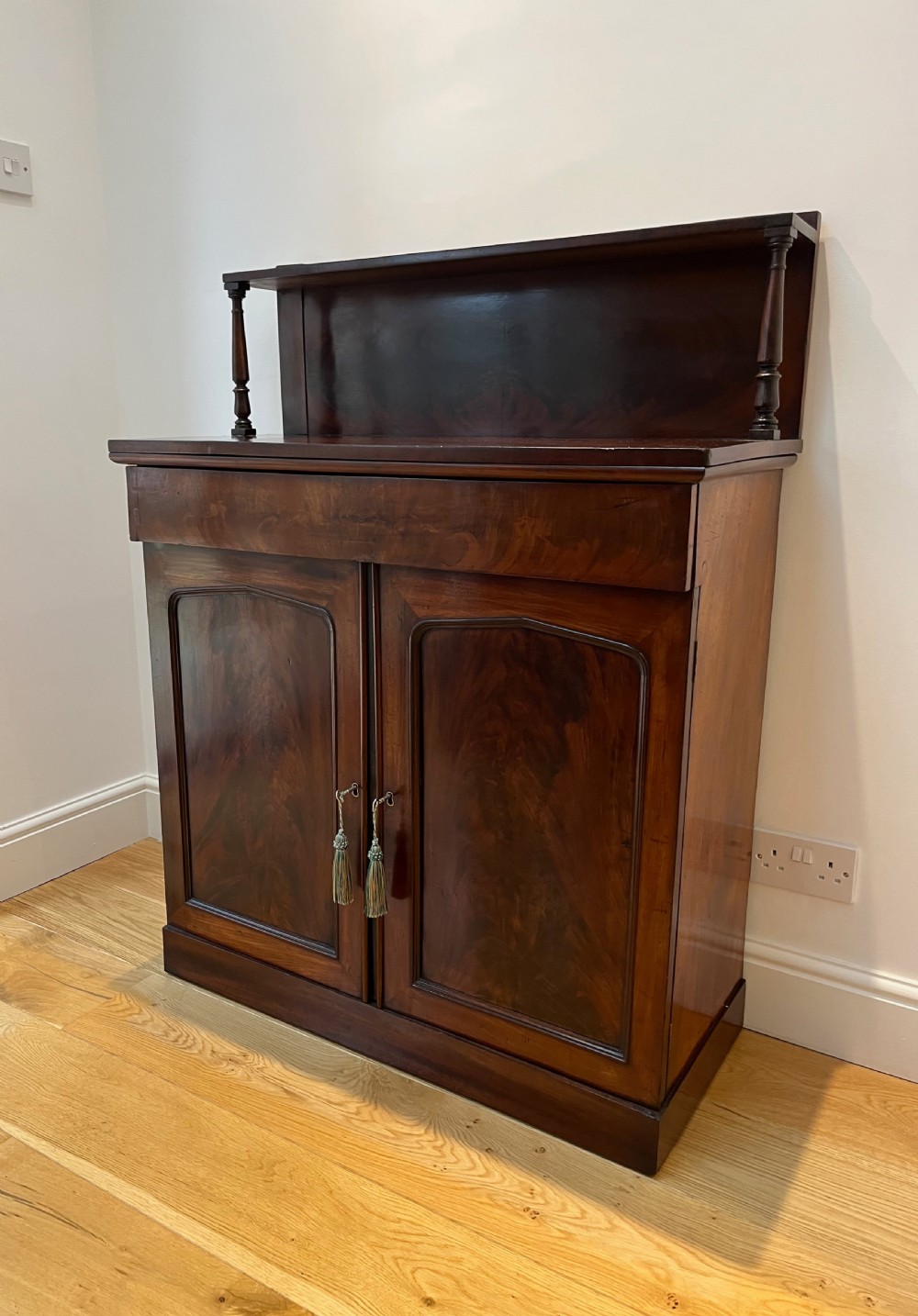 A Regency flame mahogany chiffonier cabinet featuring a top display shelf supported by turned columns over a frieze drawer and two panelled doors. View V