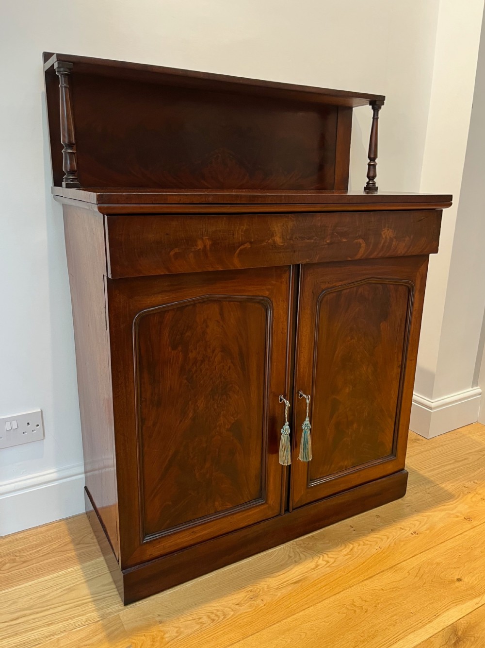 A Regency flame mahogany chiffonier cabinet featuring a top display shelf supported by turned columns over a frieze drawer and two panelled doors. View VI