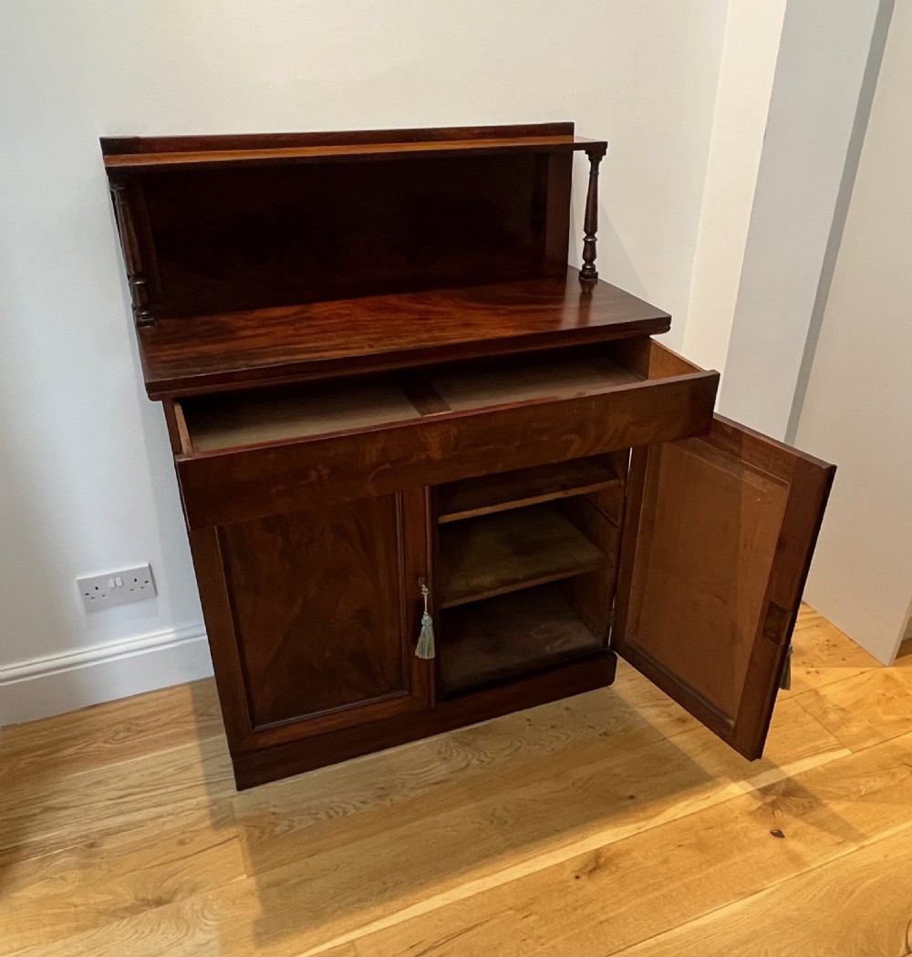 A Regency flame mahogany chiffonier cabinet featuring a top display shelf supported by turned columns over a frieze drawer and two panelled doors. View IX
