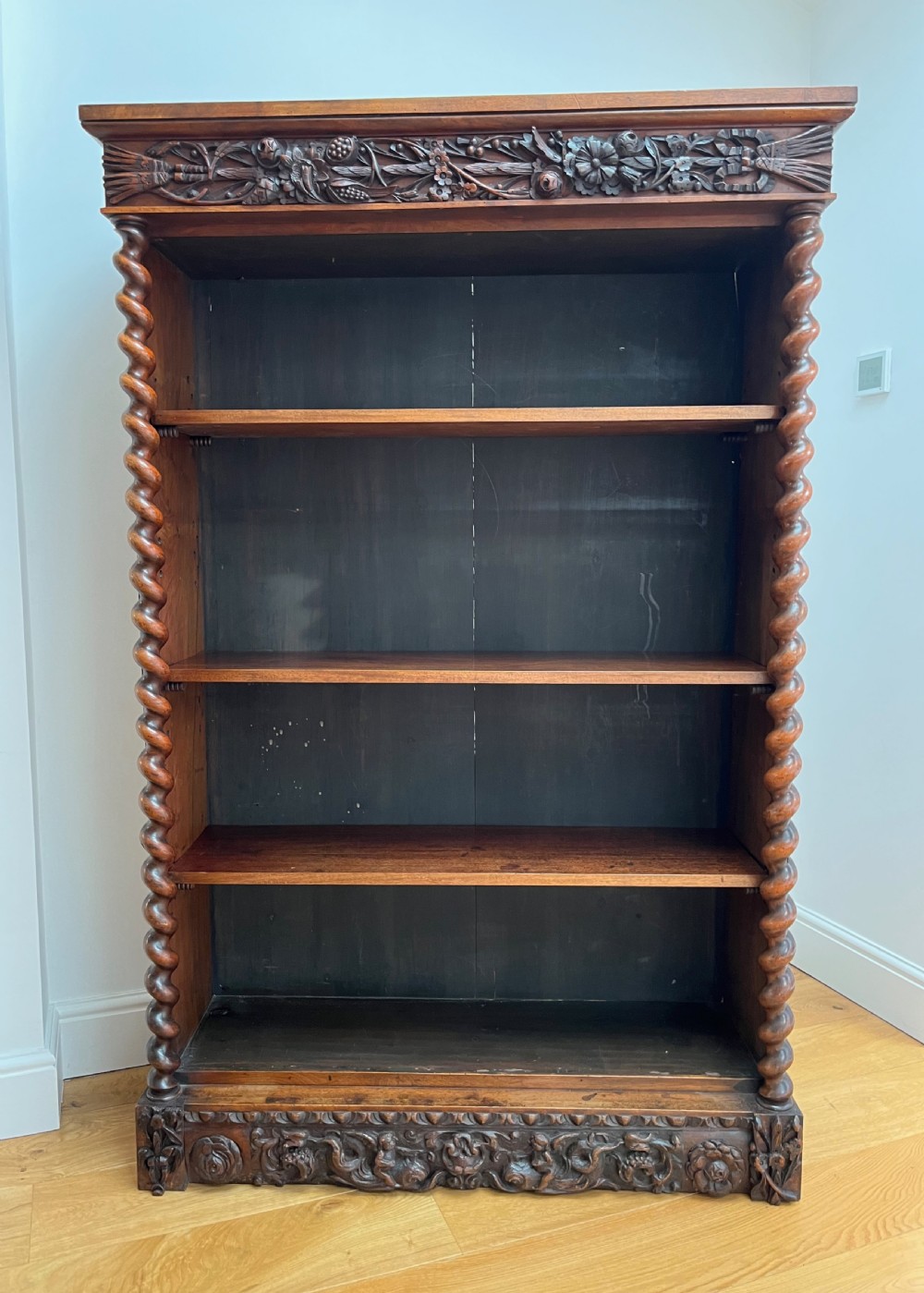 An antique dark walnut open bookcase featuring detailed foliate carved friezes at the top and base flanked by twin barley twist columns framing four shelves.
