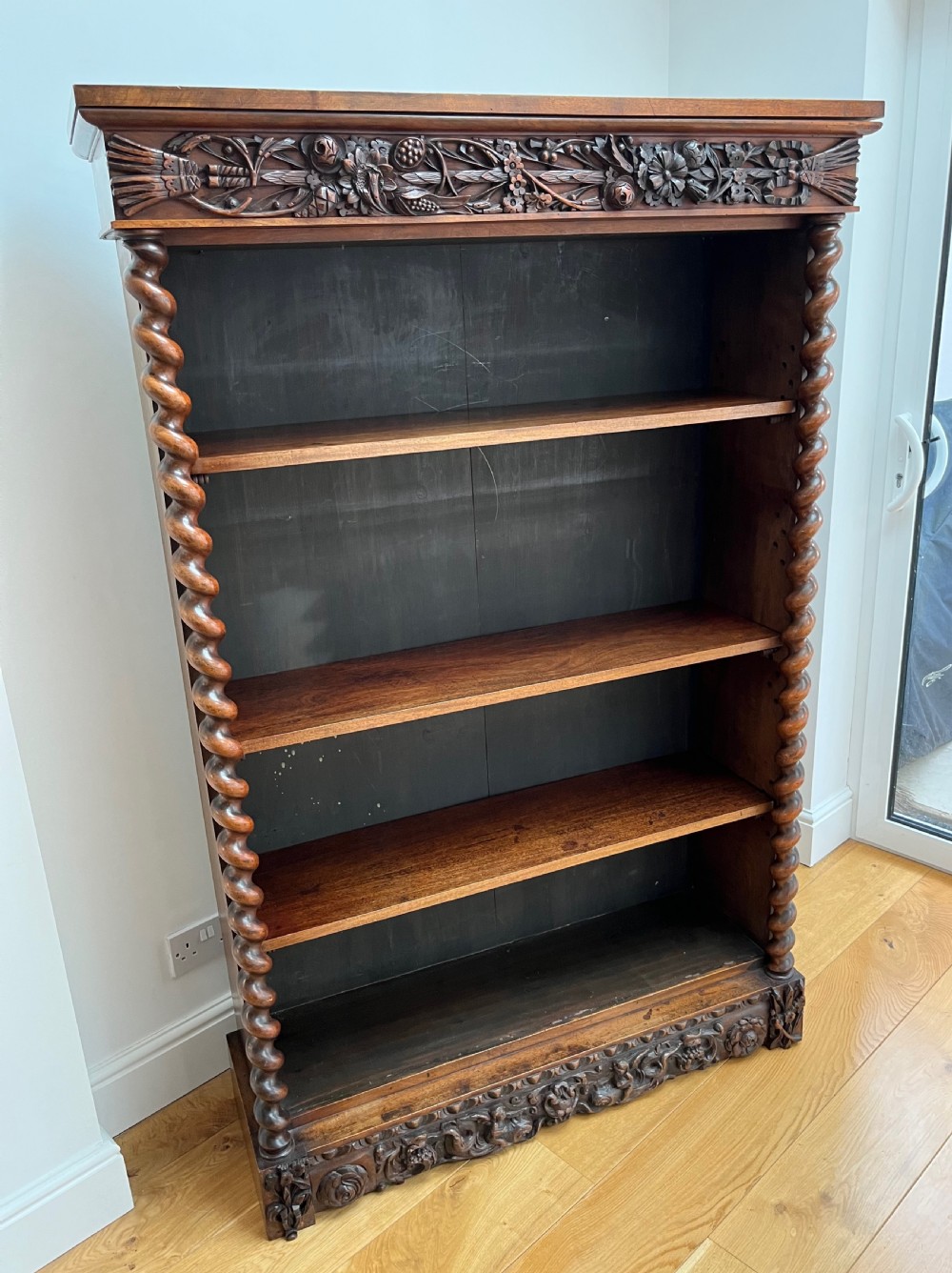 An antique dark walnut open bookcase featuring detailed foliate carved friezes at the top and base flanked by twin barley twist columns framing four shelves. View II