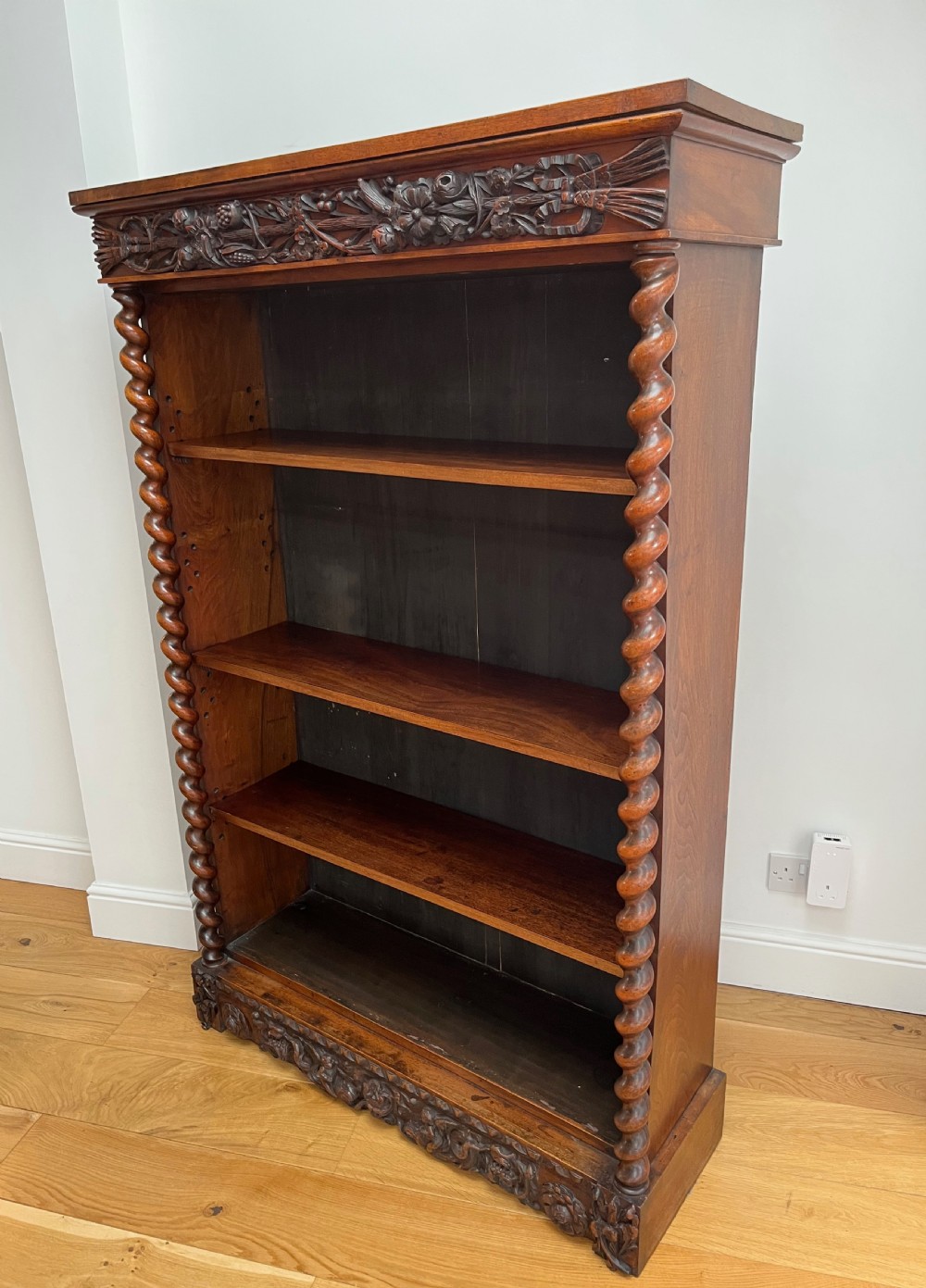 An antique dark walnut open bookcase featuring detailed foliate carved friezes at the top and base flanked by twin barley twist columns framing four shelves. View III