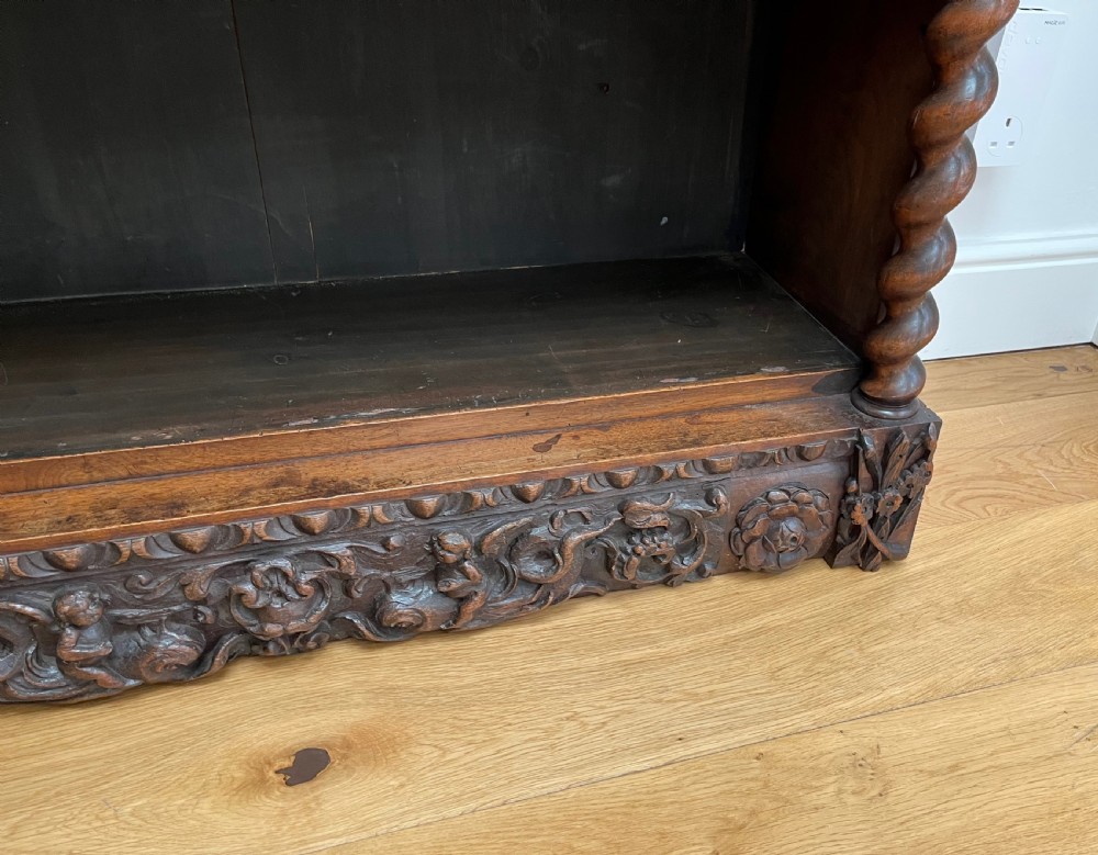 An antique dark walnut open bookcase featuring detailed foliate carved friezes at the top and base flanked by twin barley twist columns framing four shelves. View X