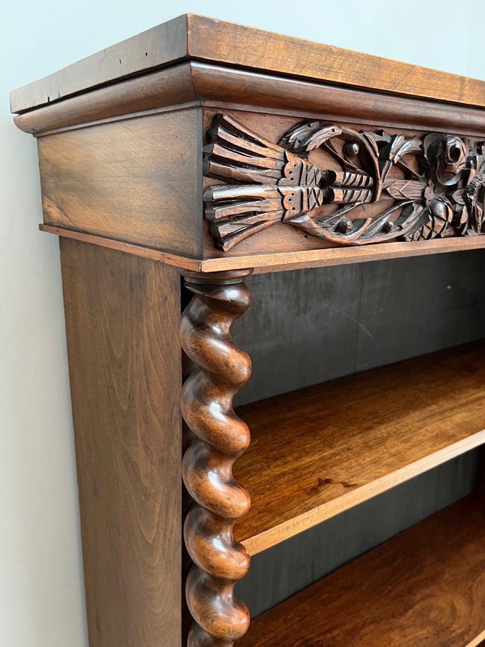 An antique dark walnut open bookcase featuring detailed foliate carved friezes at the top and base flanked by twin barley twist columns framing four shelves. View XIV