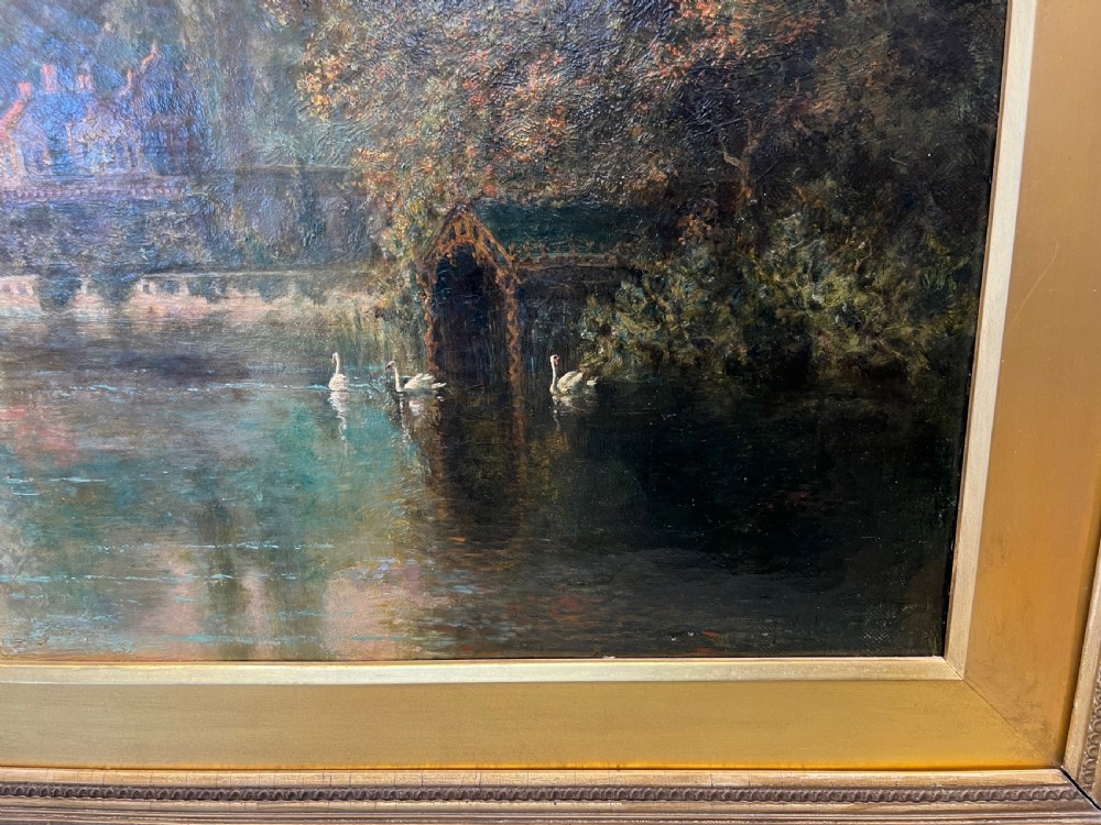 An antique framed oil painting depicting a tranquil river Thames scene with a figure in a boat, swans near a boathouse, and a riverbank estate beneath a glowing sky. View V