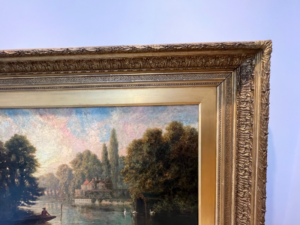 An antique framed oil painting depicting a tranquil river Thames scene with a figure in a boat, swans near a boathouse, and a riverbank estate beneath a glowing sky. View IX