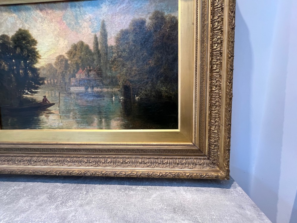 An antique framed oil painting depicting a tranquil river Thames scene with a figure in a boat, swans near a boathouse, and a riverbank estate beneath a glowing sky. View X