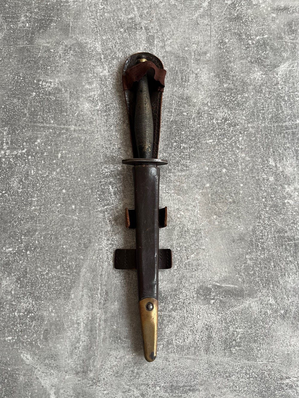 A Second World War Pattern 2 Fairbairn-Sykes combat dagger with a knurled grip resting next to its dark brown leather scabbard. View II