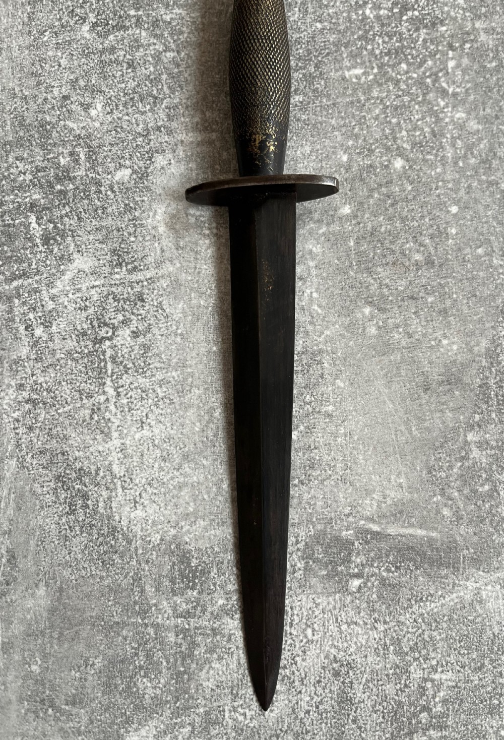A Second World War Pattern 2 Fairbairn-Sykes combat dagger with a knurled grip resting next to its dark brown leather scabbard. View III