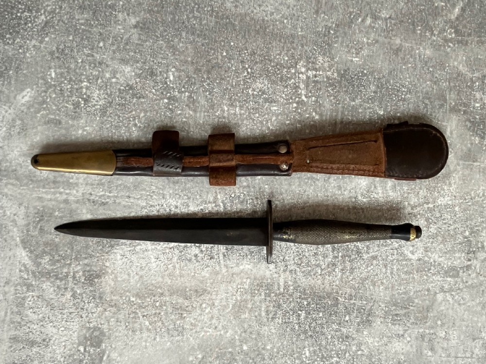 A Second World War Pattern 2 Fairbairn-Sykes combat dagger with a knurled grip resting next to its dark brown leather scabbard. View VI