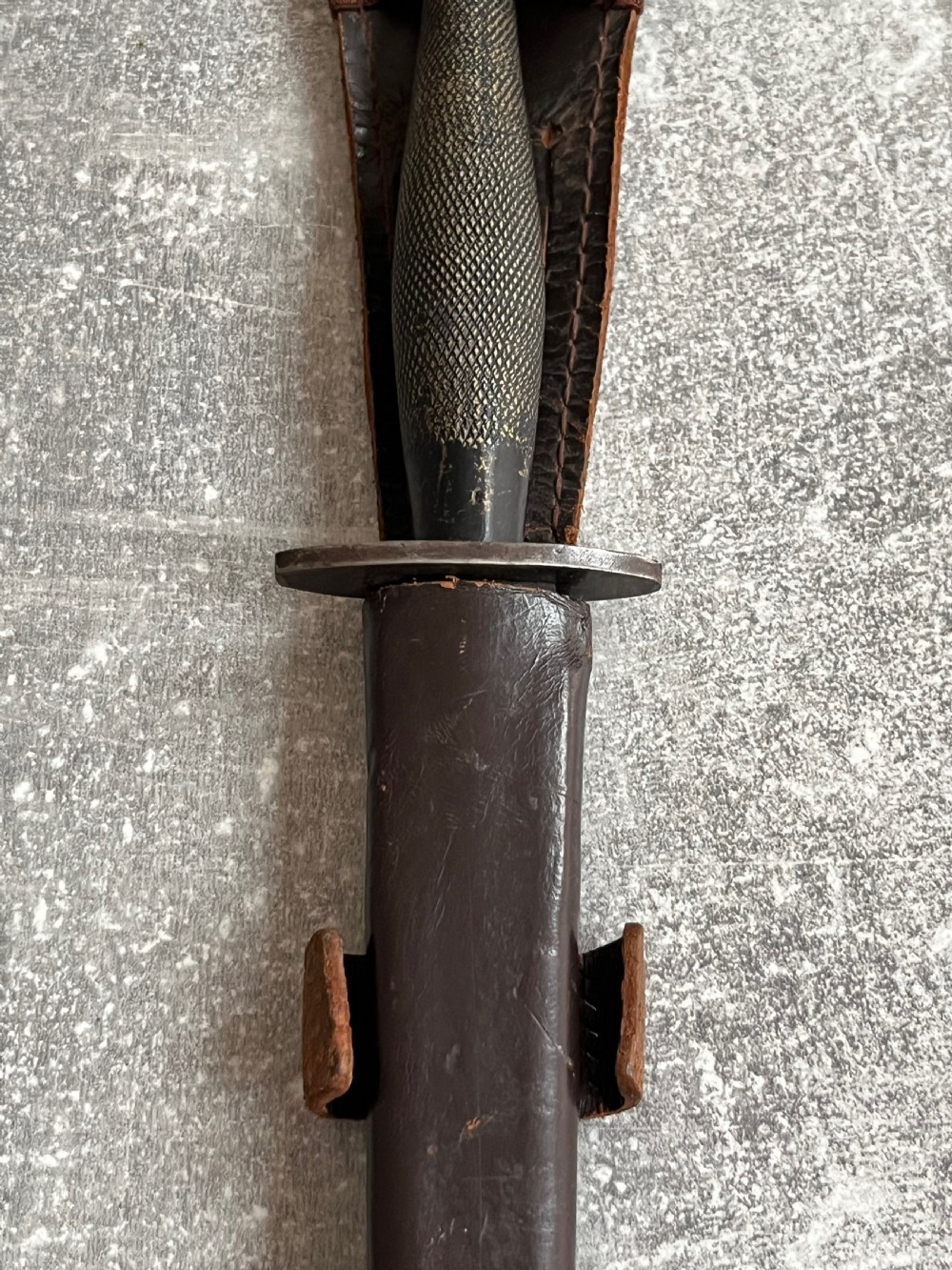 A Second World War Pattern 2 Fairbairn-Sykes combat dagger with a knurled grip resting next to its dark brown leather scabbard. View XI