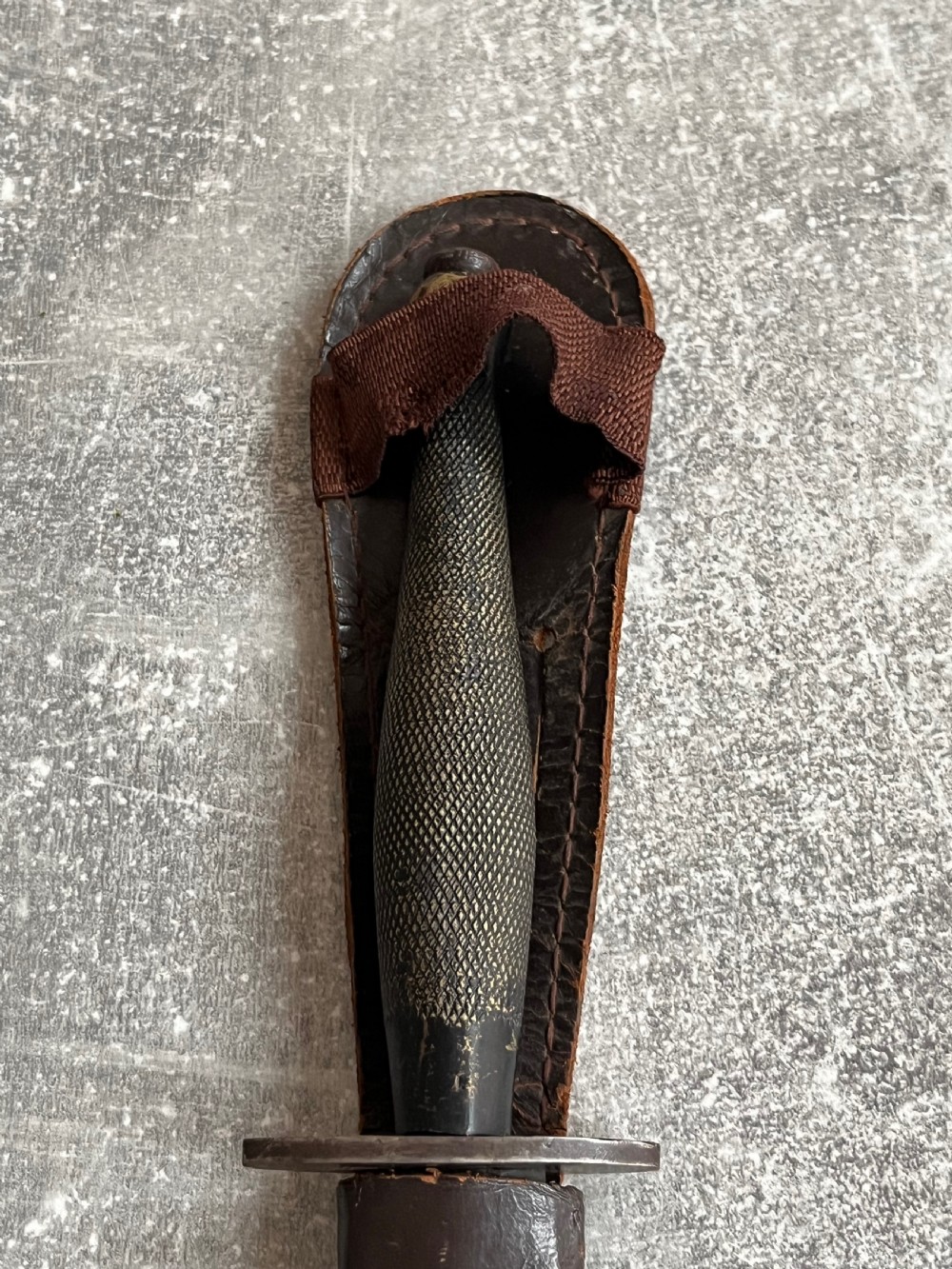 A Second World War Pattern 2 Fairbairn-Sykes combat dagger with a knurled grip resting next to its dark brown leather scabbard. View XII