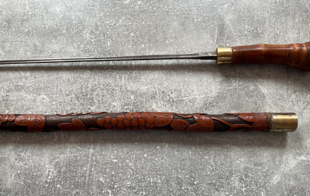 An antique East Asian carved bamboo sword stick shown unsheathed to reveal a long slender steel blade alongside its carved wooden shaft with brass fittings. View VI