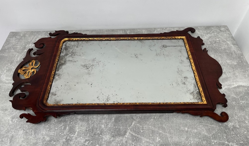 An upright rectangular wall mirror framed in dark mahogany with scroll-cut fretwork borders and a gilded carved motif at the top crest. View X