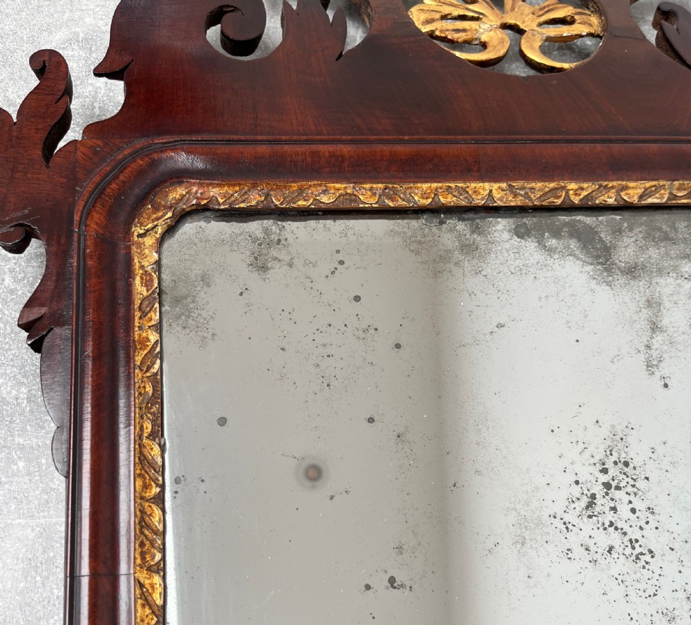 An upright rectangular wall mirror framed in dark mahogany with scroll-cut fretwork borders and a gilded carved motif at the top crest. View XII
