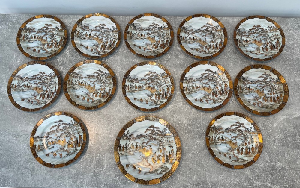 A set of twelve circular Japanese Satsuma earthenware plates featuring hand-painted landscape scenes with figures framed by wide gilded decorative borders.
