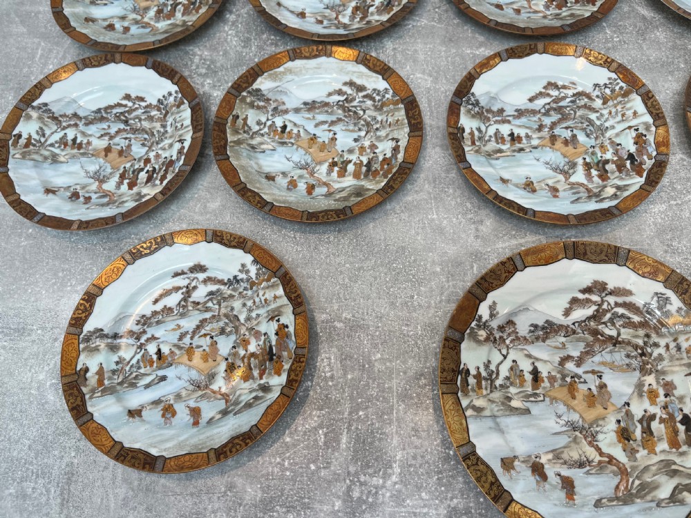 A set of twelve circular Japanese Satsuma earthenware plates featuring hand-painted landscape scenes with figures framed by wide gilded decorative borders. View V