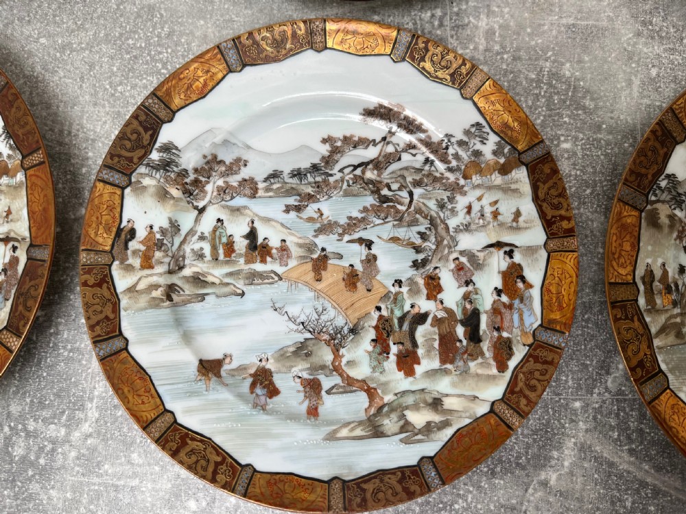 A set of twelve circular Japanese Satsuma earthenware plates featuring hand-painted landscape scenes with figures framed by wide gilded decorative borders. View IX