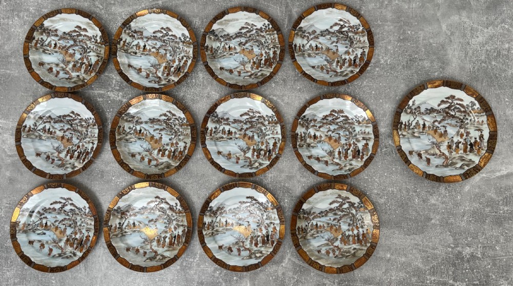 A set of twelve circular Japanese Satsuma earthenware plates featuring hand-painted landscape scenes with figures framed by wide gilded decorative borders. View X