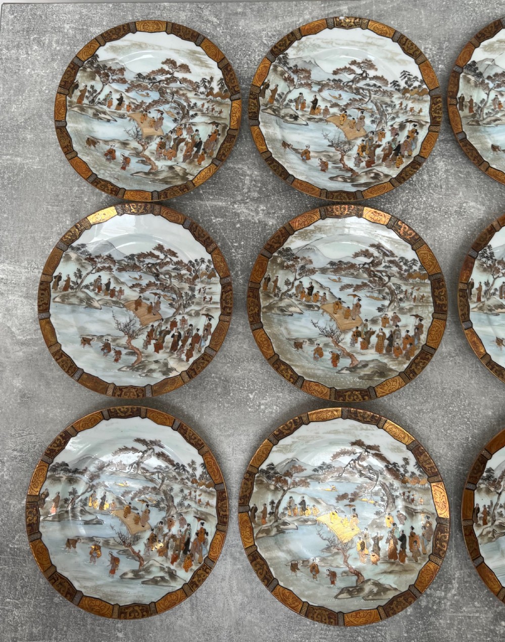 A set of twelve circular Japanese Satsuma earthenware plates featuring hand-painted landscape scenes with figures framed by wide gilded decorative borders. View XI