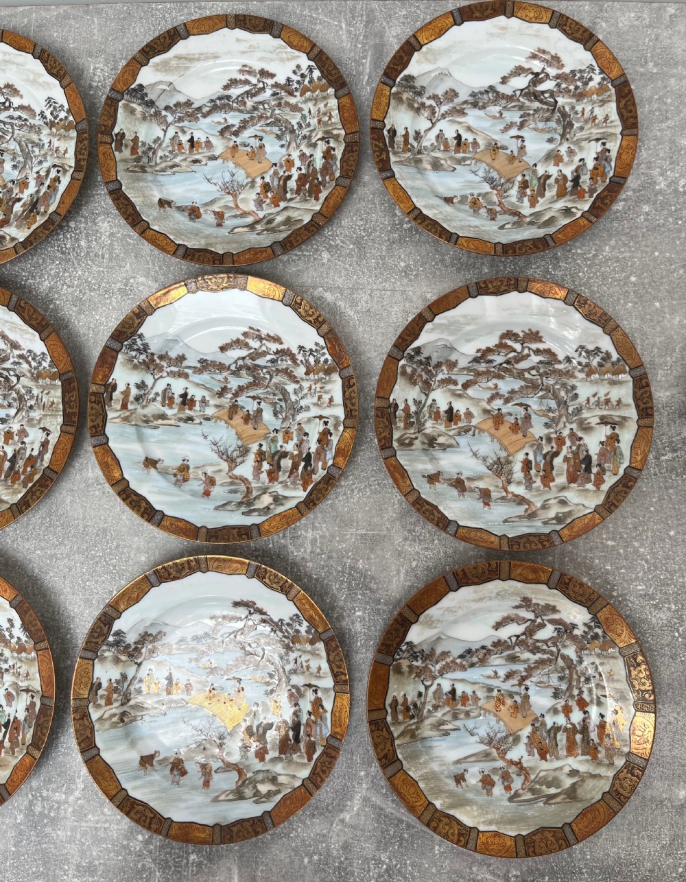 A set of twelve circular Japanese Satsuma earthenware plates featuring hand-painted landscape scenes with figures framed by wide gilded decorative borders. View XII