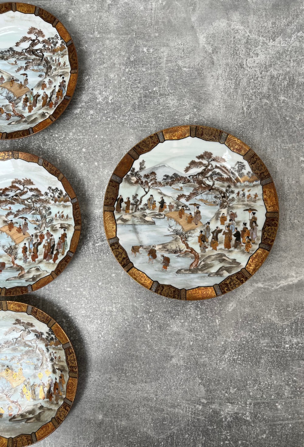 A set of twelve circular Japanese Satsuma earthenware plates featuring hand-painted landscape scenes with figures framed by wide gilded decorative borders. View XIII