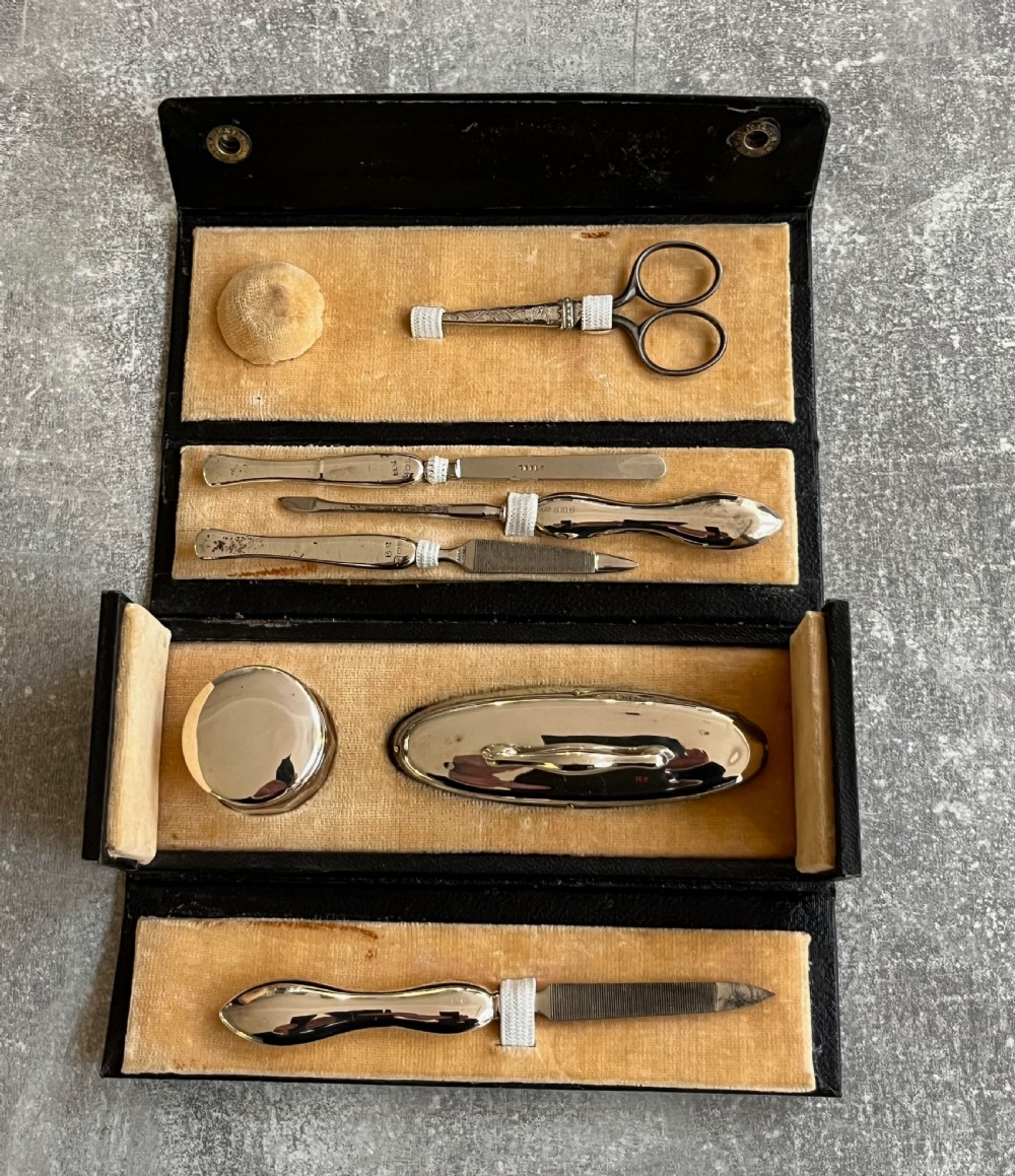 An open black folding leather case with velvet lining displaying a multi-piece antique silver manicure set including scissors, files, a nail buffer, and a small silver container.