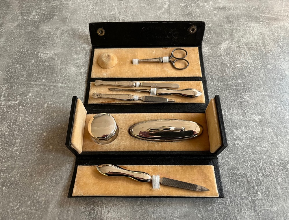 An open black folding leather case with velvet lining displaying a multi-piece antique silver manicure set including scissors, files, a nail buffer, and a small silver container. View III