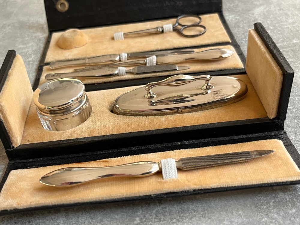 An open black folding leather case with velvet lining displaying a multi-piece antique silver manicure set including scissors, files, a nail buffer, and a small silver container. View IV