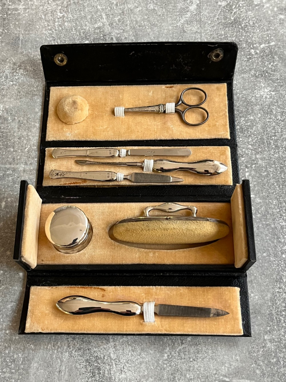 An open black folding leather case with velvet lining displaying a multi-piece antique silver manicure set including scissors, files, a nail buffer, and a small silver container. View V