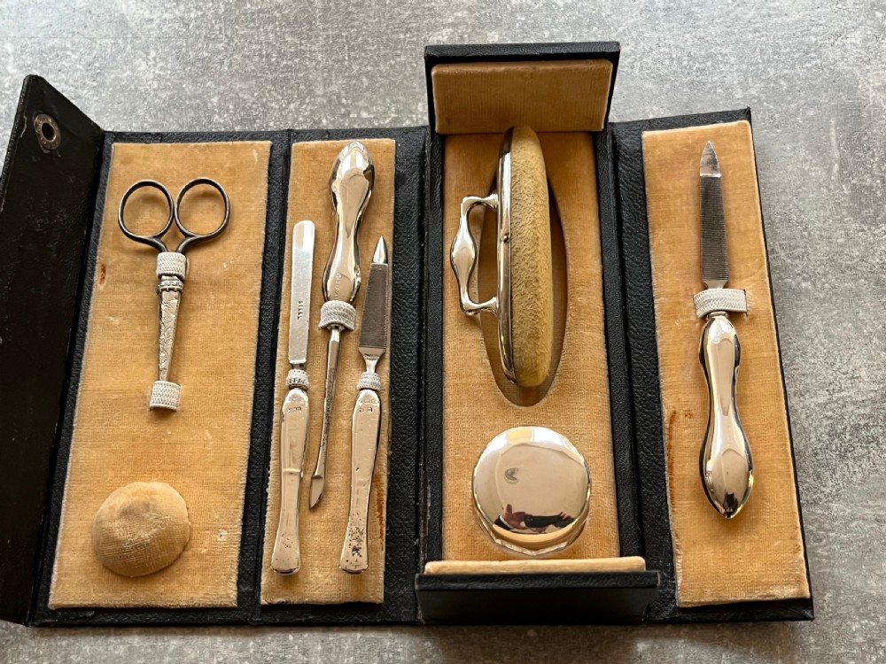 An open black folding leather case with velvet lining displaying a multi-piece antique silver manicure set including scissors, files, a nail buffer, and a small silver container. View VI