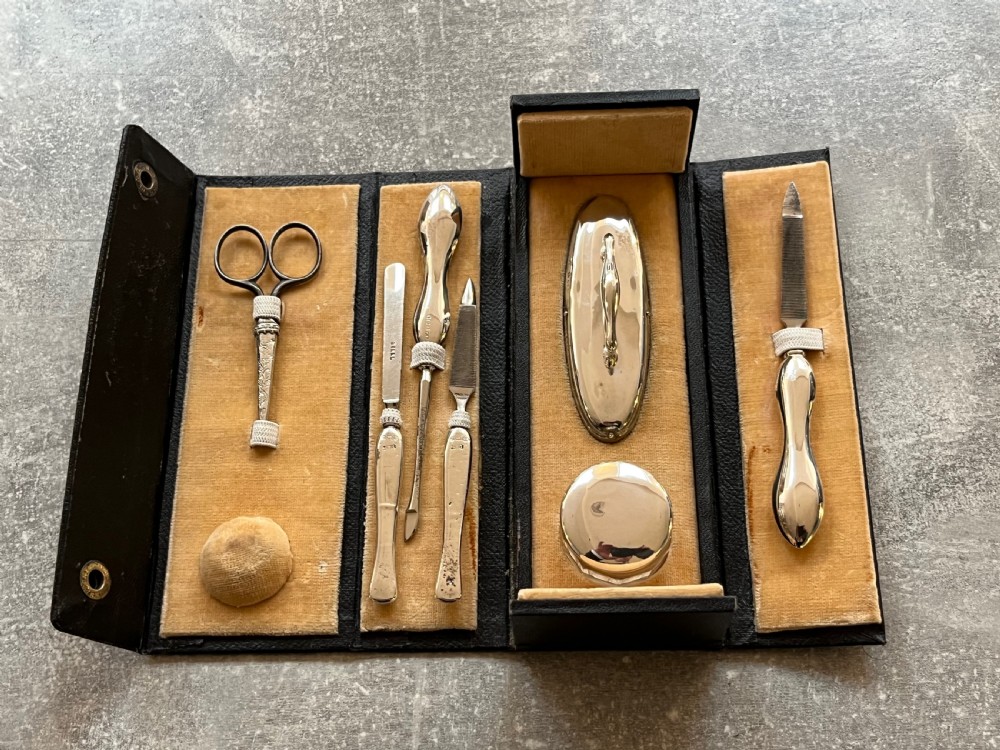 An open black folding leather case with velvet lining displaying a multi-piece antique silver manicure set including scissors, files, a nail buffer, and a small silver container. View VII