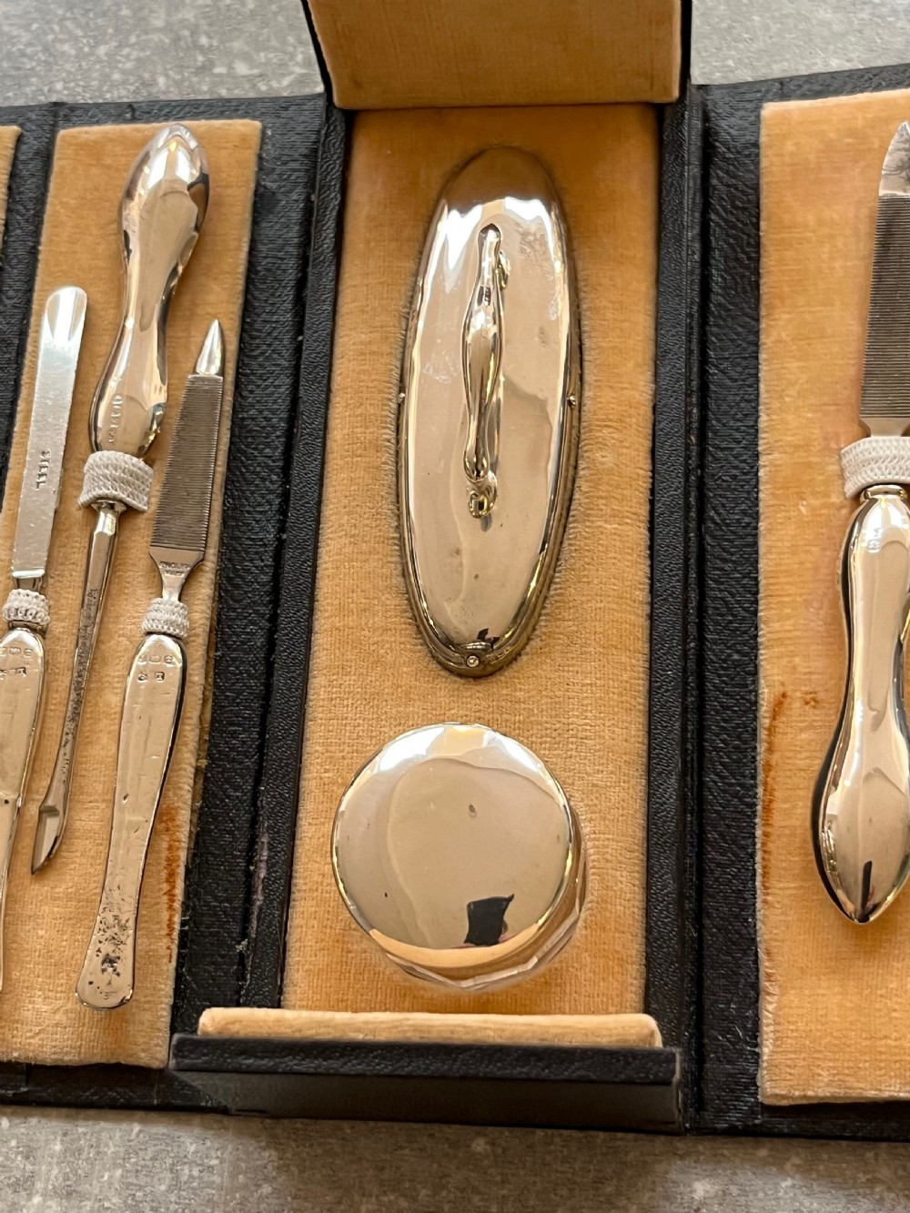 An open black folding leather case with velvet lining displaying a multi-piece antique silver manicure set including scissors, files, a nail buffer, and a small silver container. View X