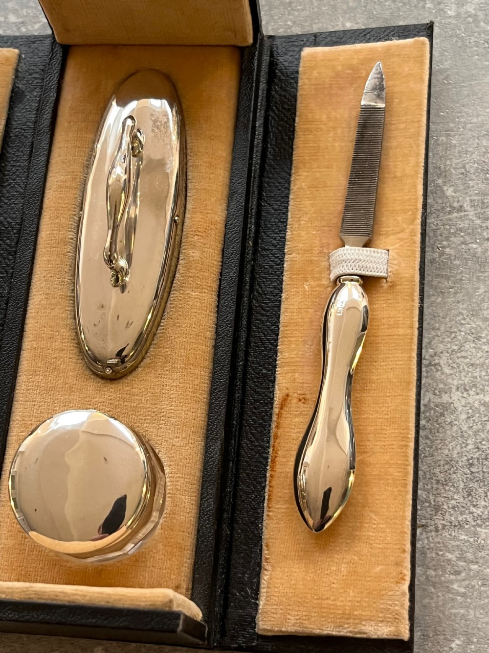 An open black folding leather case with velvet lining displaying a multi-piece antique silver manicure set including scissors, files, a nail buffer, and a small silver container. View XI