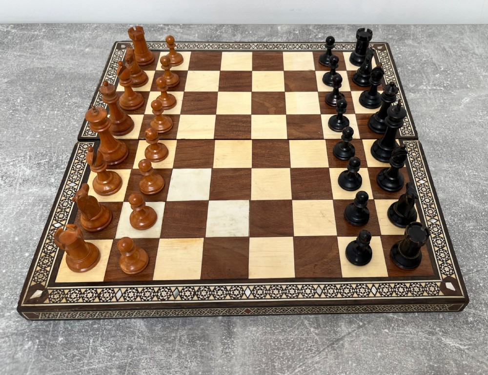 A wooden folding chess board featuring geometric inlaid borders and a full set of light and dark Staunton style chess pieces.