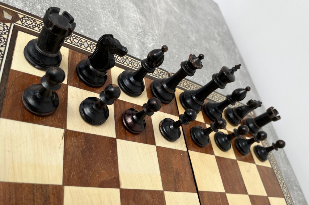 A wooden folding chess board featuring geometric inlaid borders and a full set of light and dark Staunton style chess pieces. View III